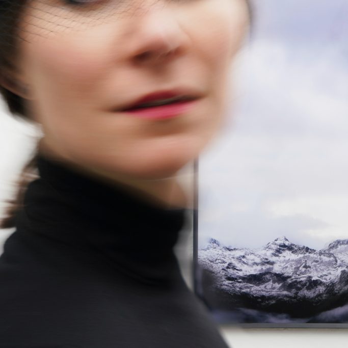 Eine elegante Frau, in ihrer Bewegung unscharf aufgenommen, vor der Fotografie „Dramatisch schön“ mit Bergpanorama und dramatischen Wolken.